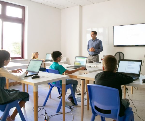 Confident teacher explaining lesson to pupils. Multiethnic children sitting at table in classroom, listening middle-aged man and using laptop computers. Childhood and digital education concept